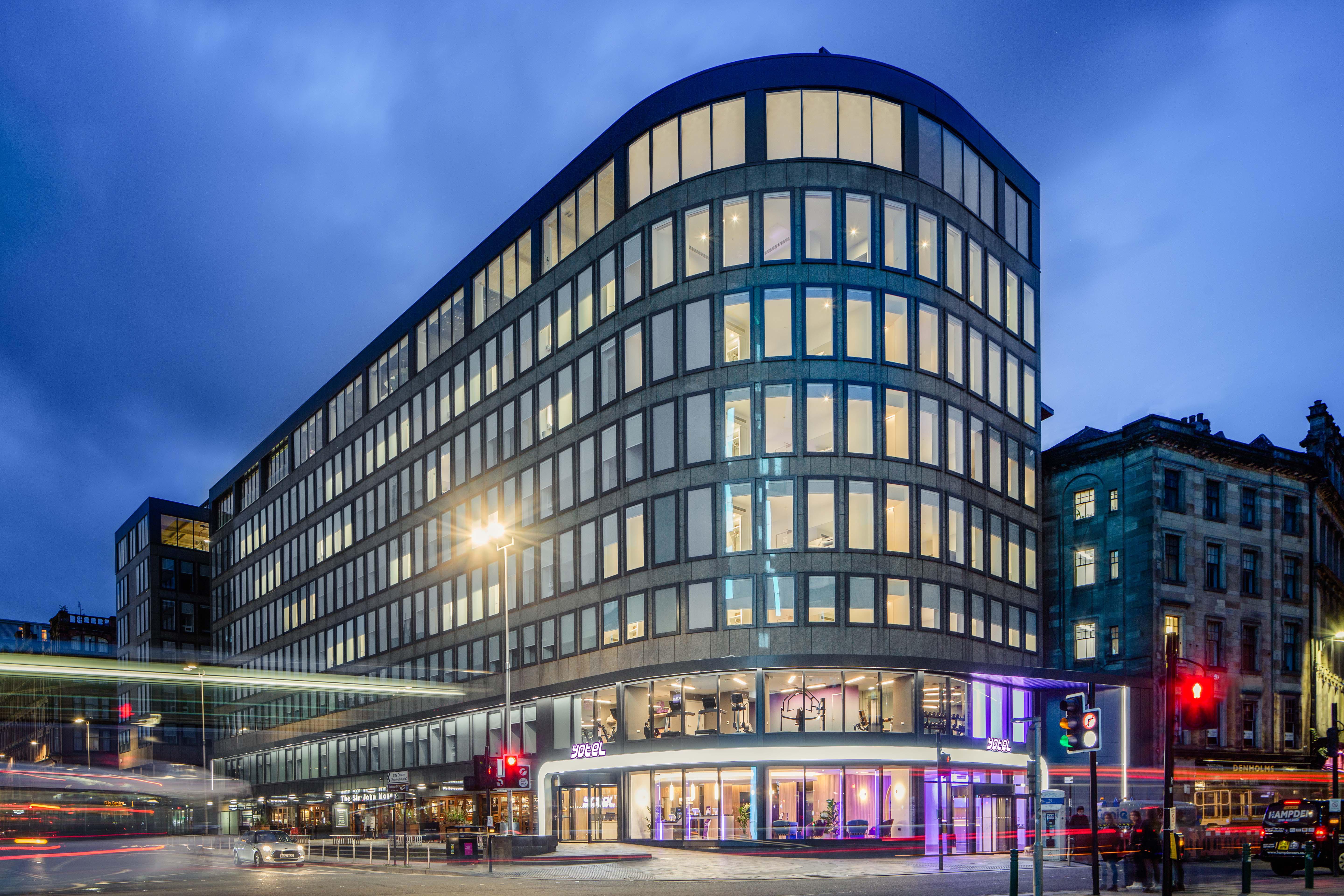Yotel 글라스고 외부 사진 The photo shows a modern office building with a distinctive curved shape and a facade made of large glass windows. It is illuminated against a twilight sky, giving it a contemporary appearance. The ground floor features large glass entrance areas, li